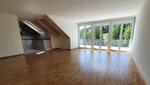 Spacious open-plan living area with hardwood flooring, large windows providing ample natural light, and access to a balcony or terrace overlooking a green outdoor area. The kitchen is integrated into the living space and features modern appliances.