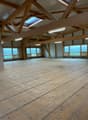 Large open space with wooden beams and trusses, hardwood flooring, and large windows providing natural light and a view of the surrounding landscape.