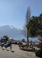 Outdoor parking lot with many cars, a lake, mountains, and a restaurant with outdoor seating.