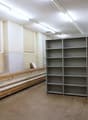 The image shows a large storage room with shelving units lining the walls. The room has a concrete floor and fluorescent lighting overhead. There are wooden benches or platforms along one wall, likely used for storage or work. The room appears to be a uti