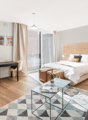 Open-plan studio apartment with hardwood floors, large windows, a bed, a sofa, a coffee table, and a desk area. The room has a modern, minimalist design with neutral colors and natural wood accents.