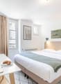 Spacious bedroom with large windows, beige curtains, white walls, hardwood flooring, and modern furniture including a bed, desk, and shelving unit. The room also features artwork on the walls and a laptop on the desk, suggesting it may be used as a worksp
