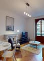 white painted walls, white hardwood floors, a fireplace, a large glass door with a window, a round wooden coffee table, a rug, a grey sectional sofa, a dining area with a round white table and chairs, a TV with a TV stand, a side table with a bowl, a lamp