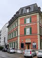 A multi-story hotel with red brick walls and green shutters, named Hotel Alpenblick, located on a street with parked cars and trees, next to another white building with many windows.