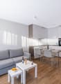 Open space living room with kitchen, dining area, and seating area with a gray couch, white tables, wooden floor, white kitchen cabinets, and stainless steel countertops.