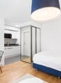 The image shows a modern and minimalist studio apartment. The room features a white and blue color scheme, with a large bed, a desk with a chair, and a kitchenette area with a microwave and other appliances. The room has hardwood floors and a large window