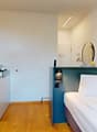 Compact kitchen with sink, faucet, coffee machine, and toaster. Bedroom with a bed, bedside table with lamp, and wardrobe. Modern bathroom with white tiles and wooden floor.