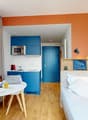 small studio, wooden floor, open plan, blue cupboards, microwave, blue door, single bed, pillows, table with chair, large window with view