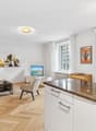 open living space with kitchen, large couch with cushions, armchair, coffee table, tv stand with flat screen tv, floor lamp, wooden parquet flooring, kitchen island with granite countertop, dining area with dining table and chairs, picture frames on the w