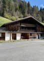 A traditional wooden chalet-style building with a sloped roof, surrounded by a grassy area and trees. The building has a covered porch and several garage doors or parking spaces on the ground floor.