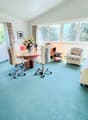 Spacious home office with large windows, wooden desk, office chairs, floral armchair, and a sofa. The room has a blue carpeted floor and appears to have access to a balcony or terrace.