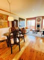 Wooden flooring, round dining table with chairs, hanging lamp, paintings on the wall, glass sliding doors, couch with pillows, side table with lamp, coffee table, rug on the floor, cabinet