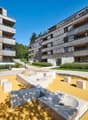 Multi-story apartment buildings with wooden facades, balconies, and a landscaped courtyard with a modern sculpture and seating area.