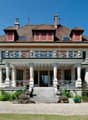 Raised ground floor, tiled roof, brown brick, white exterior, large balcony with railings, staircases, stone porch, windows, plants