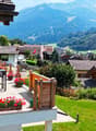 Apartment with balcony, mountain view, wooden railing, outdoor seating with table and chairs, flower pots, green lawn, several buildings and a mountain range in the background.