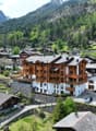 The image shows a picturesque alpine village nestled in a lush, mountainous landscape. The buildings are primarily made of wood and stone, with traditional alpine architecture featuring sloped roofs and balconies. The village is surrounded by dense forest
