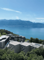 Modern building complex with multiple buildings, solar panels, surrounded by trees, overlooking a lake and mountains