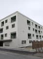 A multi-story apartment building with a modern, minimalist design. The building has several balconies and large windows. The exterior is painted in a light gray color. There are cars parked in front of the building, and the surrounding area appears to be