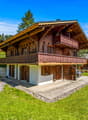 2-story wooden chalet-style house with a balcony, surrounded by a grassy lawn and pine trees in a mountainous, forested area