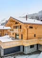 2-story wooden chalet-style house with a stone base, large balconies, and a garage. The house is surrounded by snow-covered mountains and trees.