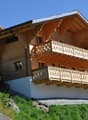 2-story wooden chalet-style house with decorative balconies, located on a hillside with a grassy slope and surrounded by trees