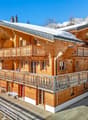 Two-story wooden chalet with a sloped roof, covered balconies, and a snowy mountain backdrop