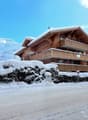 A multi-story wooden chalet-style building with snow-covered roofs and balconies, surrounded by snow-covered trees and a snowy landscape.