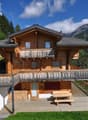 2-story wooden chalet-style house with a balcony, wooden deck, and parking space. The house is surrounded by lush green trees and mountains in the background.