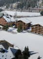 large chalet building, modern architecture, wooden balconies, snow-covered surroundings, surrounded by trees, playground
