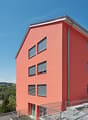 Modern apartment building, red exterior, multiple windows, metal railings, gravel pathway, nearby car