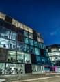 A modern, multi-story office building with a glass facade and visible parking spaces in front. The building has a sleek, contemporary design with large windows allowing natural light to enter the interior spaces.