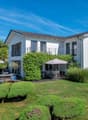 Two-story white house with balconies, surrounded by a well-maintained garden with trees and shrubs. There is a patio area with outdoor furniture in the backyard. The house has a tiled roof and large windows.