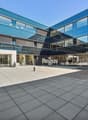 Modern glass and steel commercial building with the Regus logo prominently displayed. The building has a large open plaza area with paved flooring in front of the main entrance.