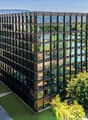 A modern, multi-story office building with a glass facade surrounded by lush greenery and landscaping. The building has a rectangular shape with numerous windows and appears to be located in an urban setting with mountains visible in the background.