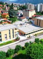 Multi-story building with a flat roof, situated near a road, surrounded by greenery and buildings