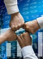 The image shows the hands of several people coming together in a fist bump, symbolizing teamwork and collaboration. The background features a cityscape with skyscrapers and financial data, suggesting a business or corporate setting.