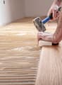 Hardwood floor being installed on concrete base, person using hammer, wall with white paint, grooves for installation