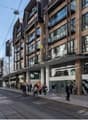 Busy city street with tram tracks, multi-story buildings with shops and restaurants, pedestrians walking on the sidewalk.