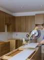 Unfinished kitchen, wooden cabinets, countertop, electrical outlet on wall, sawing machine, open door, man using mask, hammer on belt