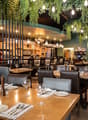 Upscale restaurant with a cozy and modern interior design. The space features wooden tables, leather chairs, and a bar area with a large selection of wines and liquors. The ceiling is adorned with hanging greenery and string lights, creating a warm and in