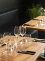 Wooden dining table set with wine glasses, cutlery, and a plant decoration. Dark walls and furniture create a cozy atmosphere. The table is set for a dining experience, with the visible features including a dishwasher, suggesting this is a restaurant sett