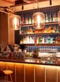 Well-stocked bar with a variety of liquor bottles, glassware, and large pendant lights hanging above the bar counter. The bar has a modern, cozy atmosphere with wooden accents and tiled walls.