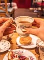 The image shows a person's hands holding a floral-patterned ceramic mug filled with a hot beverage, likely tea or coffee, on a table with other dishes and plates. The background suggests a cozy cafe or restaurant setting with wooden furniture and decor.