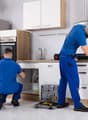 modern kitchen with stainless steel appliances, white cabinets, tiled flooring, two men in blue uniforms, toolbox and tools on the floor