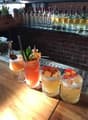 The image shows a bar setting with various cocktail glasses and bottles on display. There are three cocktail drinks on the wooden bar top, each garnished with different fruits and herbs.