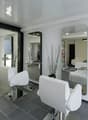 Spacious hair salon with modern, minimalist decor. White chairs and tables, black and gray tile flooring, large mirrors, and decorative branches. Appears to have a dishwasher visible in the background.
