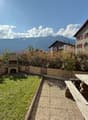 Garden: wooden table, benches, gravel path, green lawn, bushes, fireplace, wooden deck, mountains in the background.