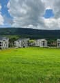 A modern residential complex with multiple apartment buildings surrounded by a large grassy field and hills in the background. The buildings have balconies and are painted in light colors.