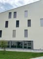 Multi-story apartment building with a modern, minimalist design. The building has large windows, balconies, and a paved outdoor area with a picnic table. The exterior is painted in a light, neutral color with black window frames and railings.