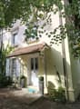 A two-story house with a yellow exterior and a brown roof. The ground floor is raised with concrete steps leading to the entrance. The windows are adorned with white shutters. The house is surrounded by a yard with trees and shrubs. There is a pathway lea